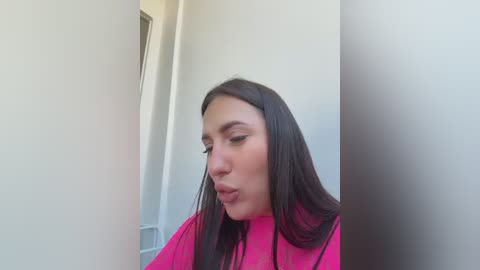 A video of a young woman with long, straight black hair, fair skin, and pink lipstick, wearing a bright pink top, looking down in a narrow, white-walled hallway.