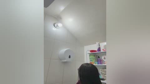 Video of a bathroom with white tiled walls and a white toilet paper holder. A woman with dark hair, facing away, is in the foreground. The room has shelves with cleaning products.