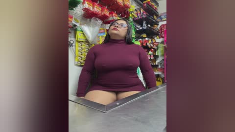 Video of a plus-sized woman with green hair and glasses, wearing a tight, burgundy mini dress, seated at a cash register in a colorful convenience store.
