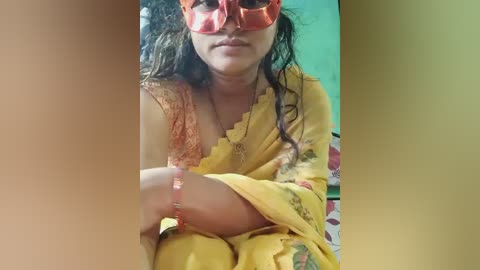 Video of a woman with light brown skin, wearing a yellow sari with floral patterns, and orange face mask, seated indoors against a green wall, with blurred background.