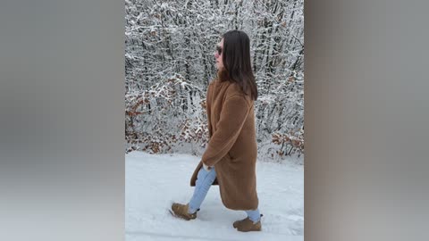 Video of a woman in a brown coat, blue jeans, and brown boots walking in a snowy forest. Snow-covered trees are in the background.