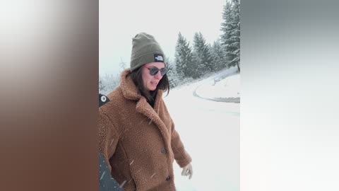 A video of a bearded man in a brown fur coat, green beanie, and dark sunglasses, standing on a snowy path with a blurred background of snow-covered trees.