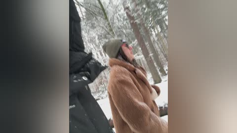 Video of a person in a beige coat, green hat, and sunglasses, looking up at a snow-covered statue in a snowy forest, blurred background.