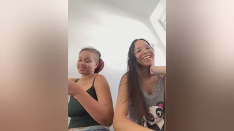 Video of two smiling women in casual attire, one wearing a green tank top and the other a gray tank top with a panda design, seated in a brightly lit room with a white ceiling.