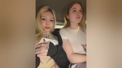 Video of two young women, one with long blonde hair and black jacket, the other with long brown hair and white shirt, sitting in a dimly lit car.