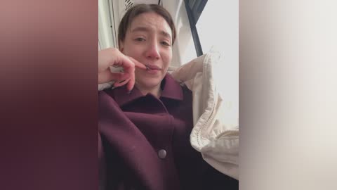 Media: Video of a Caucasian woman with light skin and brown hair, wearing a maroon coat, holding a white shirt. She appears to be inside a vehicle, with a blurred red background.