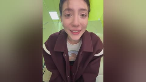 Media: A video of a young woman with light skin and brown hair, smiling, wearing a maroon jacket over a white T-shirt, in a green-tiled room with fluorescent lighting.