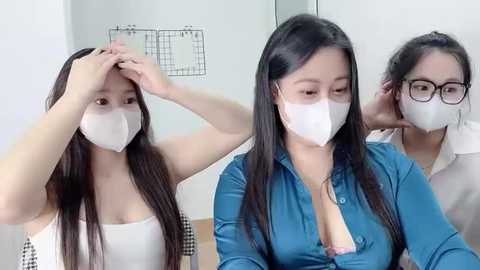 Video of three East Asian women with long black hair, wearing white face masks and blue shirts, touching their foreheads in a room with white walls and a black grid art piece.