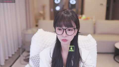 A video of an Asian woman with long black hair and glasses, wearing a white robe, sitting on a white cushioned bench in a modern, minimalistic living room.