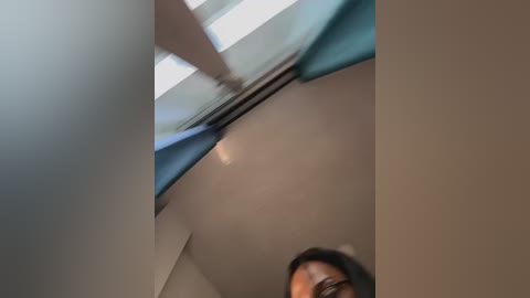 A blurred video of a person with dark hair and glasses, partially visible from the waist up, standing in a modern room with light-colored walls and a ceiling skylight.