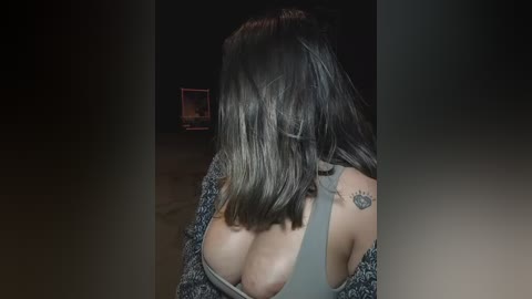 Media: Video of a woman with shoulder-length brown hair, wearing a gray off-the-shoulder top, exposing her large breasts, and a tattoo of a sun on her left shoulder. Background is dark, with a faint, blurred television screen in the distance.
