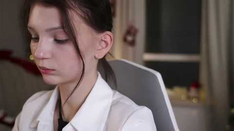 A video of a young Caucasian woman with fair skin and dark brown hair in a ponytail, wearing a white lab coat, seated indoors, with a blurred background showing a window and a bed.