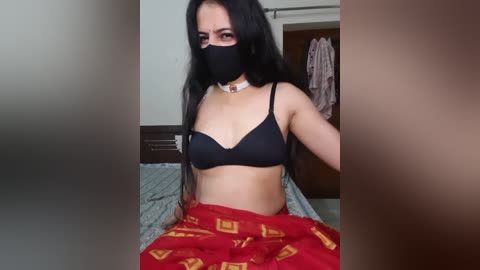 Video of a young woman with long black hair, wearing a black bra, red pajama pants with yellow patterns, and a black face mask, sitting on a bed in a simple bedroom.