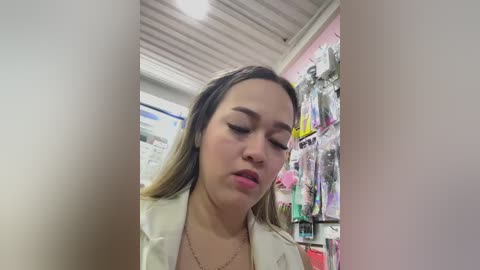 Video of a woman with light skin and long brown hair, wearing a beige jacket, looking contemplative in a store with hanging clothes and a metal ceiling.