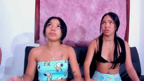Video of two Asian women with light skin, short black hair, wearing strapless tops with cartoon dogs, sitting on a black couch against a pink wall.