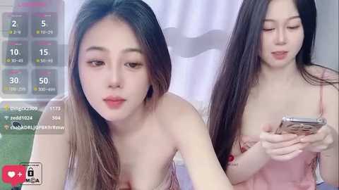 A video of two Asian women with long black hair, one in a pink dress, the other in a strapless top, both looking at a phone. Background includes a white curtain and a digital clock display.