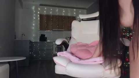 A video of a modern, dimly-lit bedroom with white, plush furniture, a woman with long, straight dark hair, and fairy lights on the wall.