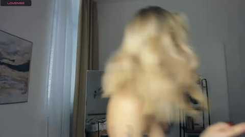 Video of a blonde woman with long hair, standing naked in a dimly lit bedroom, with a mirror reflecting her backside, and a blurred image of a male in the foreground.