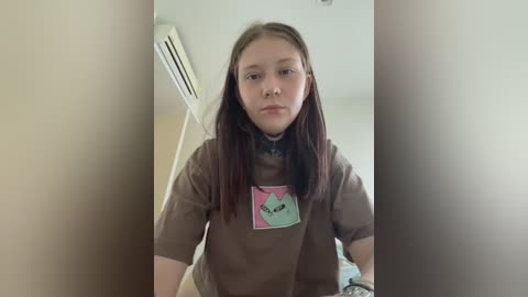 Video of a young girl with straight, shoulder-length brown hair, wearing a brown t-shirt with a pink and green heart design, standing indoors near an air conditioner.