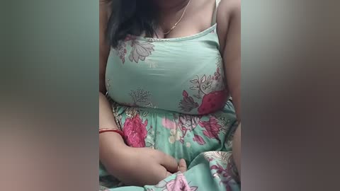 Video of a woman with medium brown skin, wearing a light turquoise floral dress with pink and red flowers, seated indoors, hands clasped, blurry background.