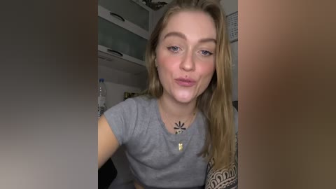 Video of a young Caucasian woman with light skin, long blonde hair, wearing a gray crop top, smiling with pouty lips. She has a tattoo sleeve on her right arm and a necklace with a star pendant. Background shows a kitchen with shelves and a water bottle.