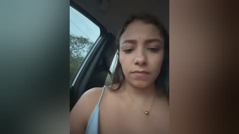 Video of a young Latina woman with light skin, straight brown hair, wearing a light blue spaghetti-strap top, inside a car, looking tired, with a blurred car door and tree outside the window.