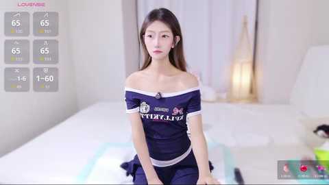 A video of an East Asian woman with long brown hair, wearing a blue off-shoulder sports jersey and matching shorts, sitting on a bed in a minimalist, white bedroom.