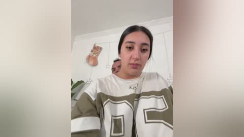 A video of a young woman with dark hair, wearing a white and green hockey jersey with the number 8, standing in a room with a white door and a hanging Christmas ornament.