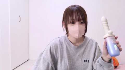 A video of a young East Asian woman with short brown hair, wearing a gray sweatshirt and a mask, holding a blue and white dildo in a sterile, white room with a closed door.