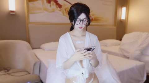 Video of a fair-skinned woman with dark hair in a white robe, holding a smartphone, in a modern, warmly lit bedroom with beige furniture and a floral wall art.