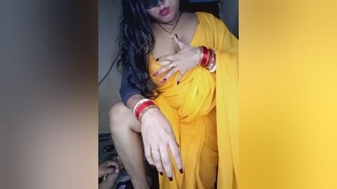 Video of a South Asian woman with medium skin tone, wearing a yellow sari, multiple bangles, and red lipstick, sitting on a bed, with a yellow wall background.