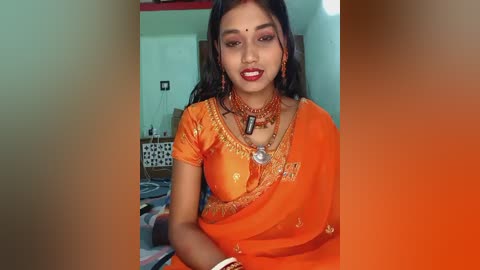 Video of a South Asian woman in an orange saree with intricate embroidery, wearing red bangles and a necklace, seated indoors with a bed and white walls in the background.