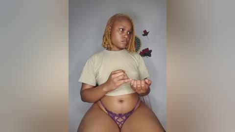 A video of a plus-size Black woman with a curvy figure, wearing a beige crop top and purple lace panties, standing against a plain white wall adorned with red and green flowers.