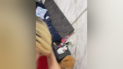 A video of a blonde woman in a red dress sitting on a bed with a grey headboard, surrounded by stuffed animals and a teddy bear.