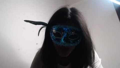 Video of a person with long, straight black hair wearing a blue, glittery masquerade mask with feathers, set against a plain, light-colored background. The person's facial expression is slightly amused.