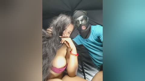 Video of a woman with long black hair, wearing a pink lace bra, and a man in a black mask, both inside a dimly lit, dark tent.