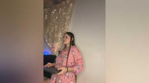 Video of a young woman with medium brown skin and long dark hair in a pink pajama set, playing a keyboard against a backdrop of twinkling fairy lights and a draped curtain.