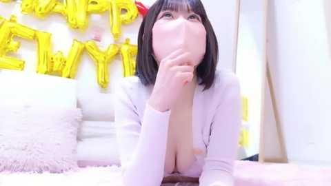 Video of an Asian woman with straight black hair and fair skin, wearing a white long-sleeve top with a deep V-neck, lying on a plush carpet. Background features a white sofa and yellow \"Happy Birthday\" balloons.