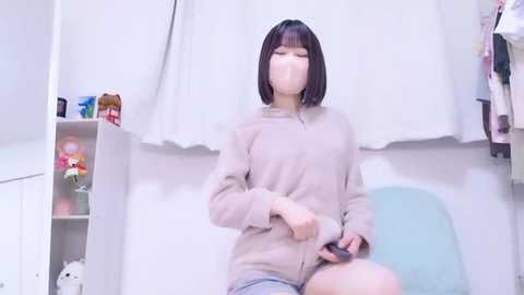 Video of a young East Asian woman with straight black hair, wearing a beige sweater and light blue shorts, sitting in a light blue chair. Background includes a white curtain, a shelf with stuffed animals, and a hanging jacket.
