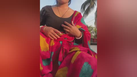 Video of a woman wearing a vibrant, multicolored saree with a black top, seated outdoors. She has medium skin tone, wearing a gold necklace and bracelets. The background features palm trees and a clear blue sky.