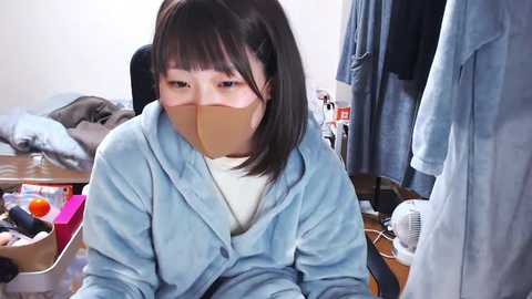 Video of a young Asian woman with straight black hair, wearing a light blue robe and beige duct tape over her mouth, sitting on a desk in a cluttered room with clothes, a fan, and a box in the background.
