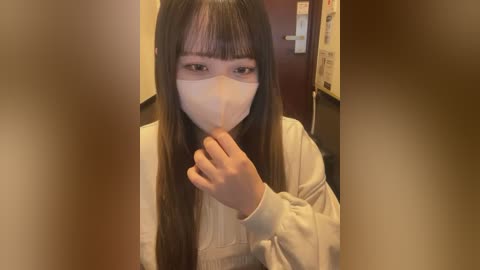 Video of an Asian woman with long black hair, wearing a white mask, holding her finger to her lips in a shushing gesture, indoors with a dark wooden door and beige walls in the background.