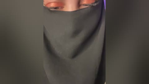Video of a person wearing a black face veil, with their nose and mouth covered, against a dark, blurred background.