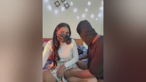 A video captures a Black woman with long hair, wearing a mask and colorful floral dress, sitting on a bed with a Black man kneeling beside her, touching her leg.