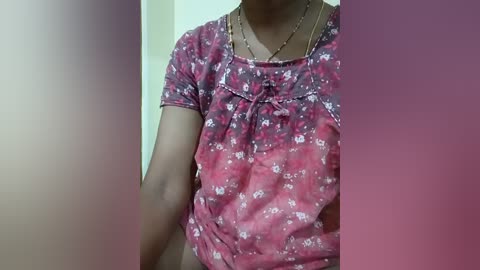 Video of a light-skinned woman wearing a floral-patterned red and white blouse, with a gold necklace. She is standing in front of a blurred background, possibly indoors.