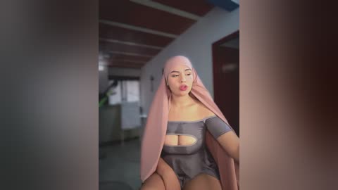 Video of a pale-skinned woman with a bald head and long pink hair, wearing a revealing, off-the-shoulder gray bodysuit, sitting in a dimly lit hallway.