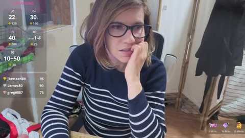 Video of a woman with short, light brown hair, wearing glasses and a navy-blue and white striped sweater, sitting at a cluttered wooden table. Background shows a messy room with clothes and a mirror.