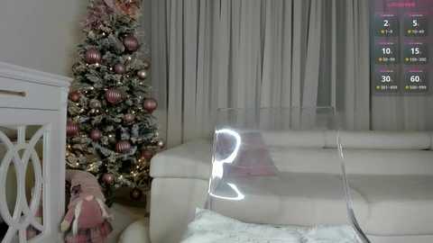 Video of a cozy living room with a Christmas tree adorned with pink and white ornaments, a white couch, and a clear acrylic coffee table. A TV screen displays a sports game.