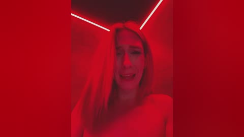 A video of a young woman with long blonde hair and a fair complexion, smiling, set against a red-lit, triangular ceiling. She wears a white top, and the background is dimly lit, creating a moody, intimate atmosphere.