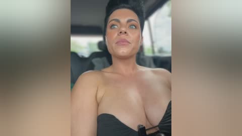 A video of a woman with light skin, large breasts, and black hair, wearing a strapless black dress, sitting in a car with blurred background.